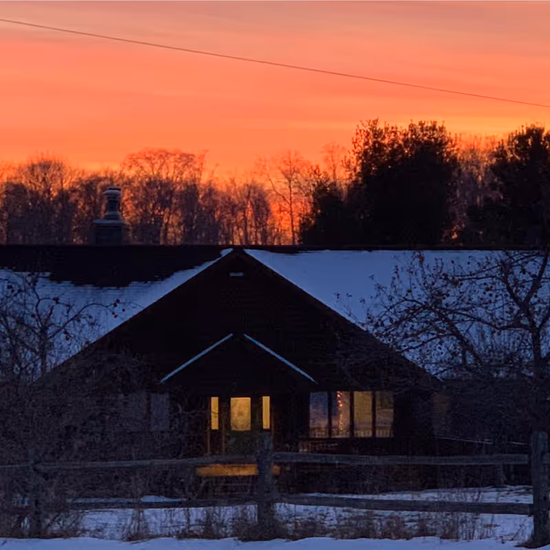 A dark building with a snow-covered roof at dusk, with warm light glowing from the windows. Bare trees and a wooden fence are visible in the foreground, and the sky is a vibrant orange and pink from the setting sun.