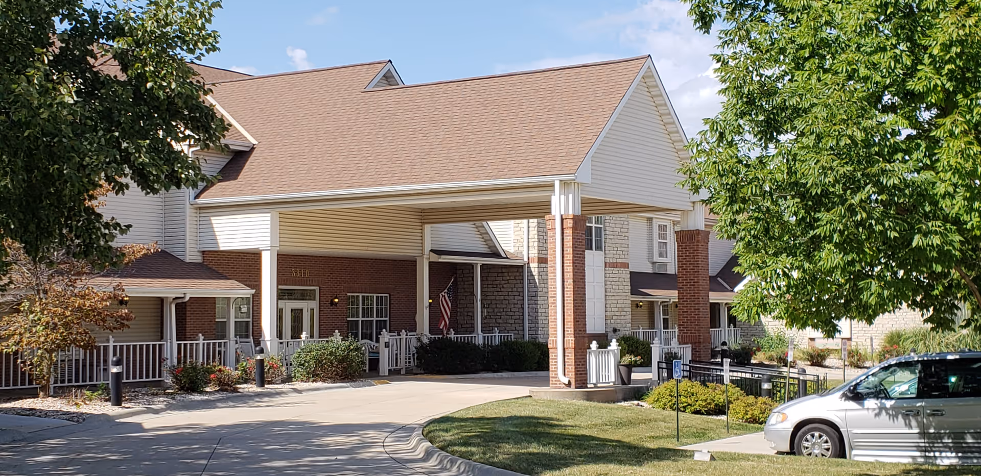 Front exterior view of a senior living facility named Villages At Vintage Oaks, featuring a covered entrance supported by brick columns, surrounded by trees and landscaping, with a silver minivan parked nearby.