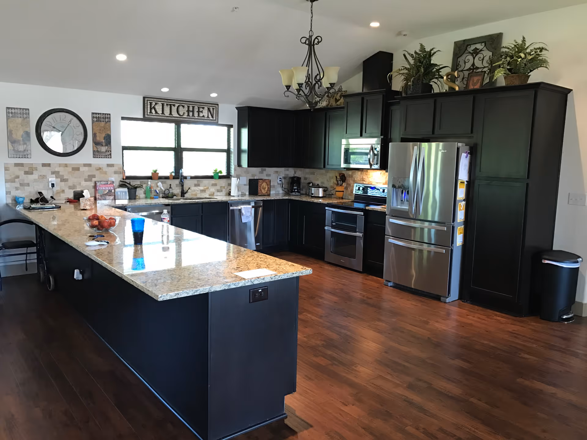 A modern kitchen with dark cabinetry and stainless steel appliances including a refrigerator, oven, and microwave. There is a large granite countertop island with a bowl of apples and a blue cup on it. The kitchen has wooden flooring, a decorative chandelier, and a sign above the window that reads 'KITCHEN'. Various plants and decorative items are placed on top of the cabinets.