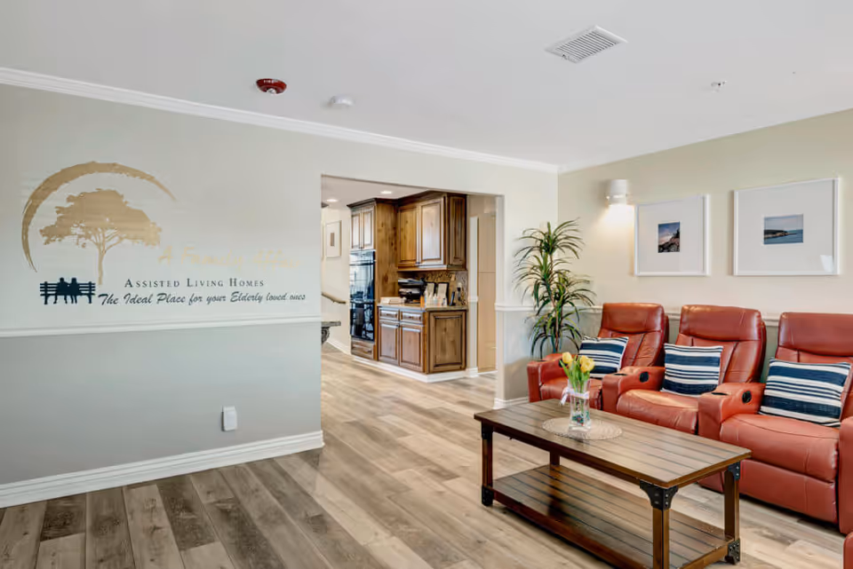 A cozy living room area with three red leather recliners, each with a blue and white striped pillow. A wooden coffee table with a glass vase holding yellow tulips is in front of the chairs. The walls are light-colored with two framed pictures and a wall-mounted light fixture. To the left, there is an open doorway leading to a kitchen area with wooden cabinets and modern appliances. The floor is wood-patterned laminate. On the left wall, there is a decorative sign with a tree logo and text about assisted living homes.