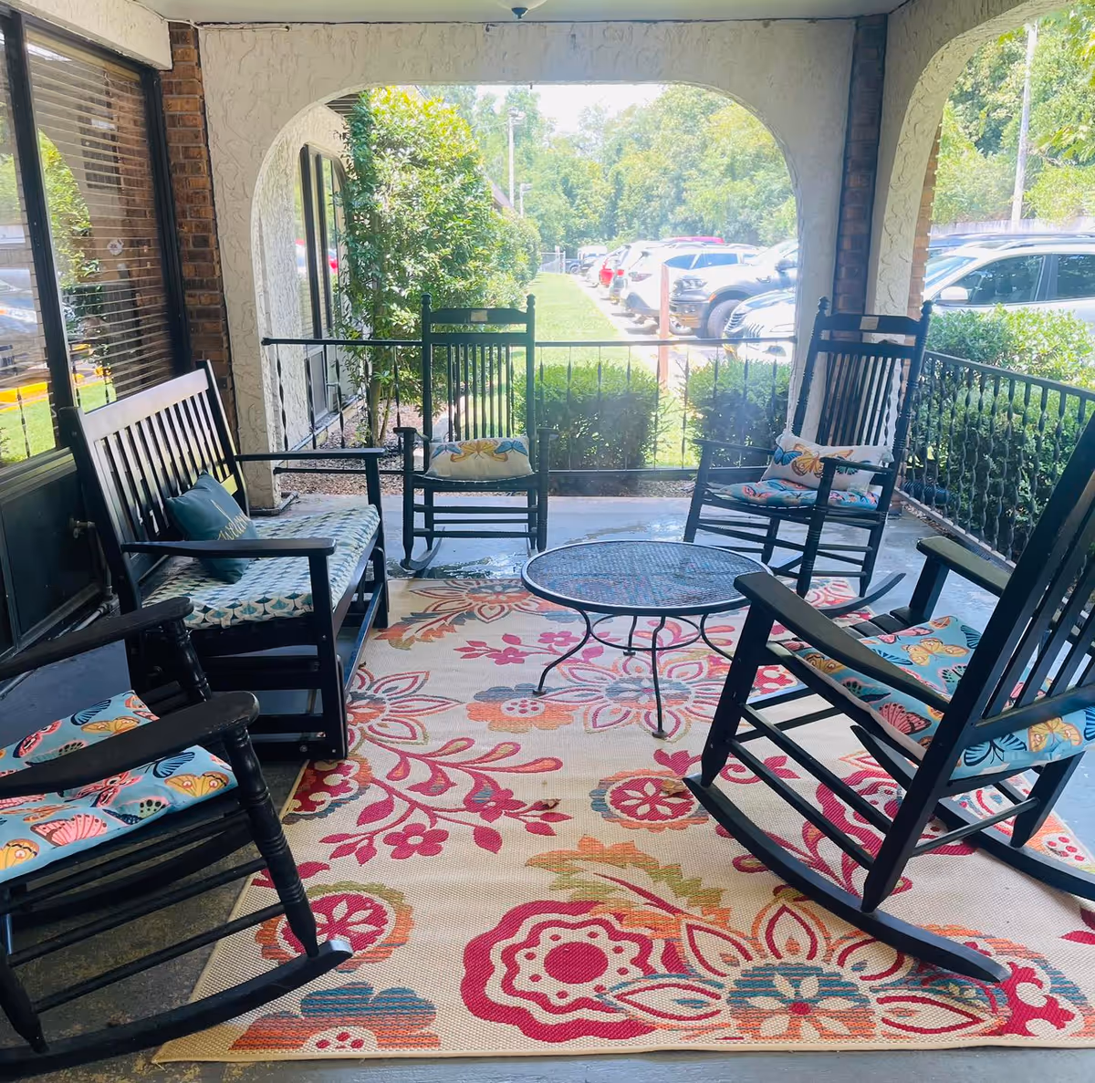 Covered outdoor patio area with black wooden rocking chairs and a bench arranged around a round metal table on a colorful floral rug. The patio overlooks a parking lot with several cars and greenery in the background.