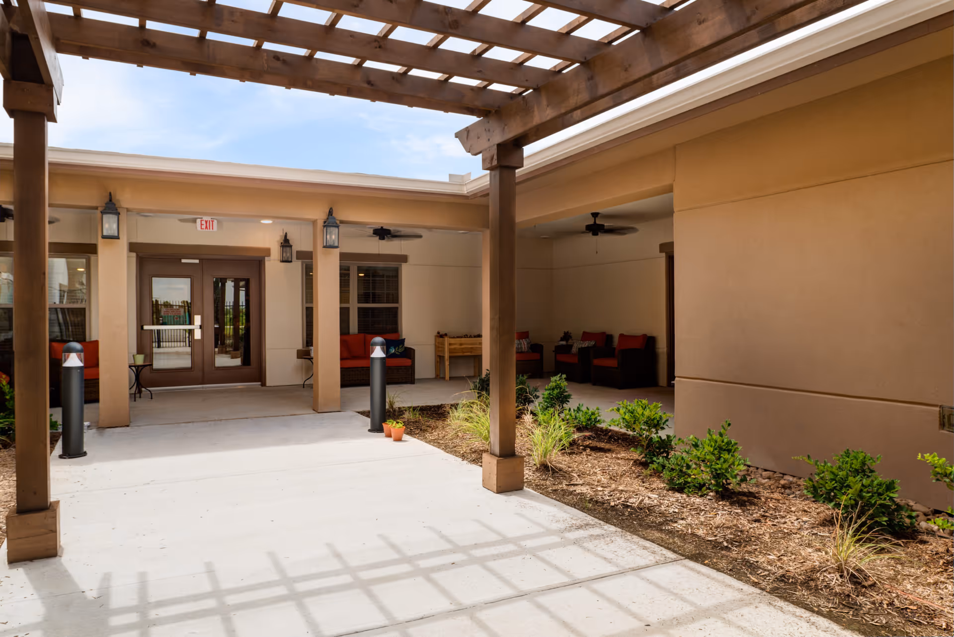 Covered outdoor entrance with a wooden pergola, walkway, landscaped beds, and seating near glass double doors.