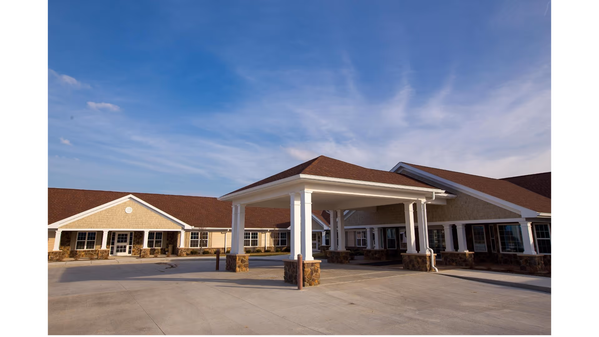 Exterior view of Meadow Grove Transitional Care facility showing a single-story building with a covered entrance supported by white columns, stone accents on the lower walls, and a red roof under a blue sky.