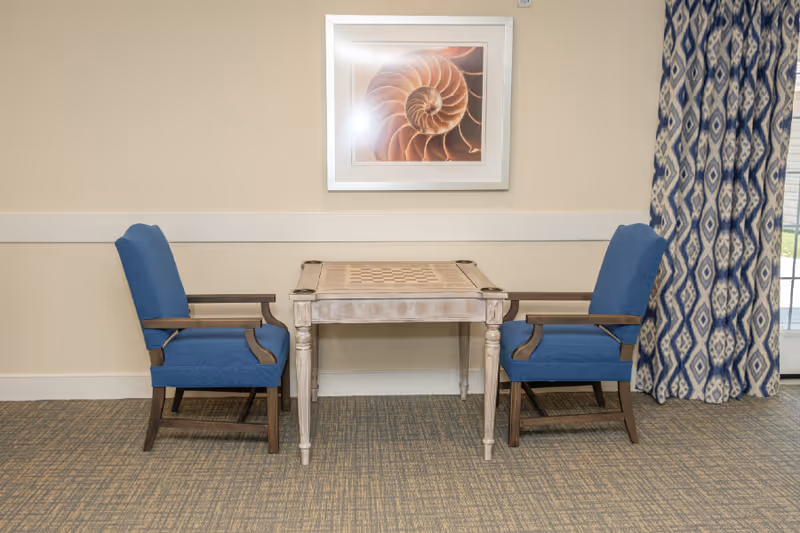 A small wooden table with a built-in chessboard is positioned between two blue upholstered armchairs with wooden frames. Behind the table on the wall is a framed picture of a nautilus shell. To the right, patterned blue and white curtains partially cover a window or glass door.