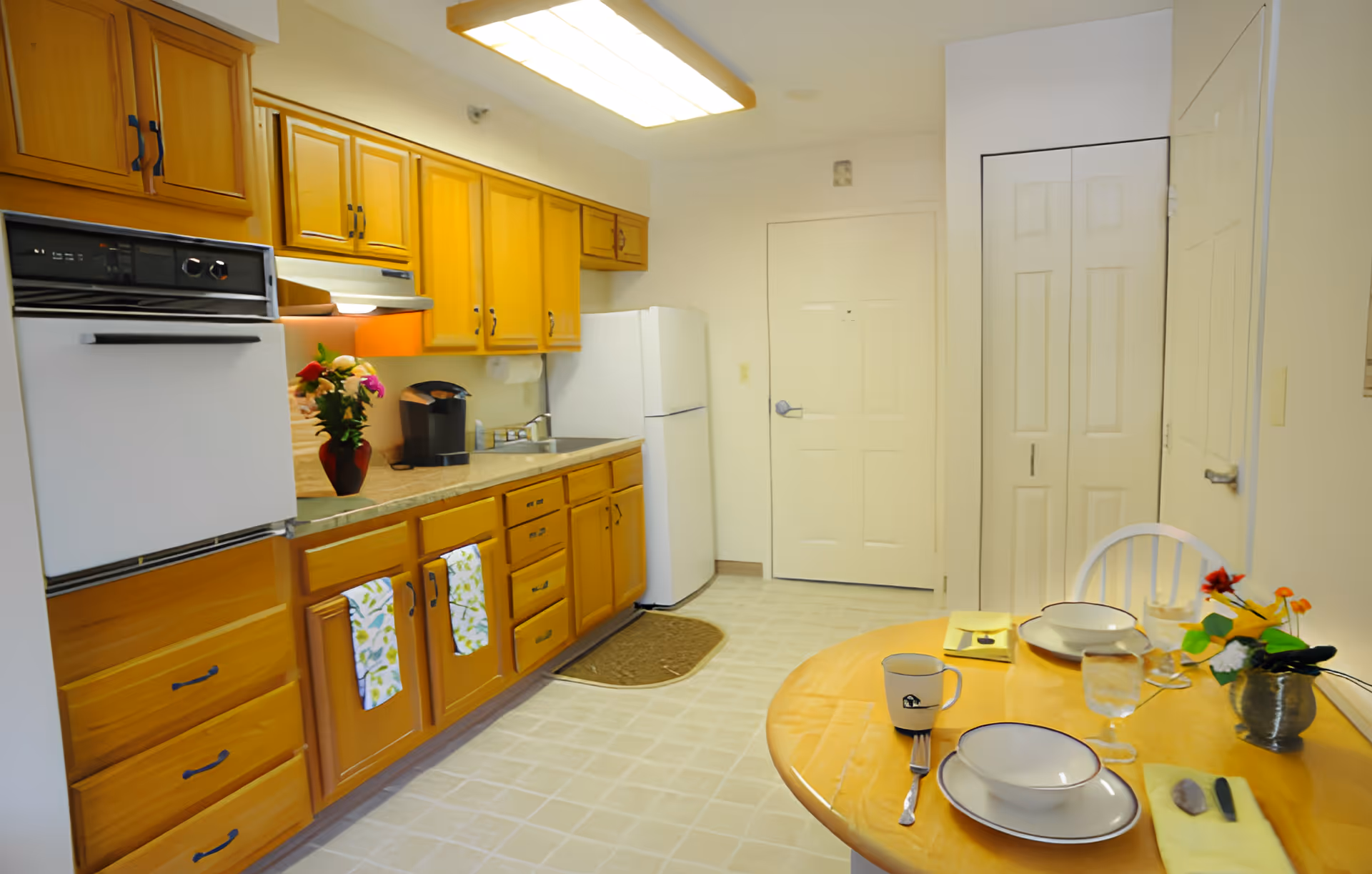 A bright kitchen with wooden cabinets, a white refrigerator, an oven, and a countertop with a coffee maker and a vase of flowers. A small round dining table is set with two place settings, including plates, glasses, utensils, and napkins, along with a small flower arrangement. The floor is tiled, and there is a door and a closet with white doors in the background.