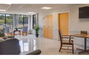 Bright communal sitting area with chairs, small tables, a potted plant, and a wall-mounted TV beside large windows.