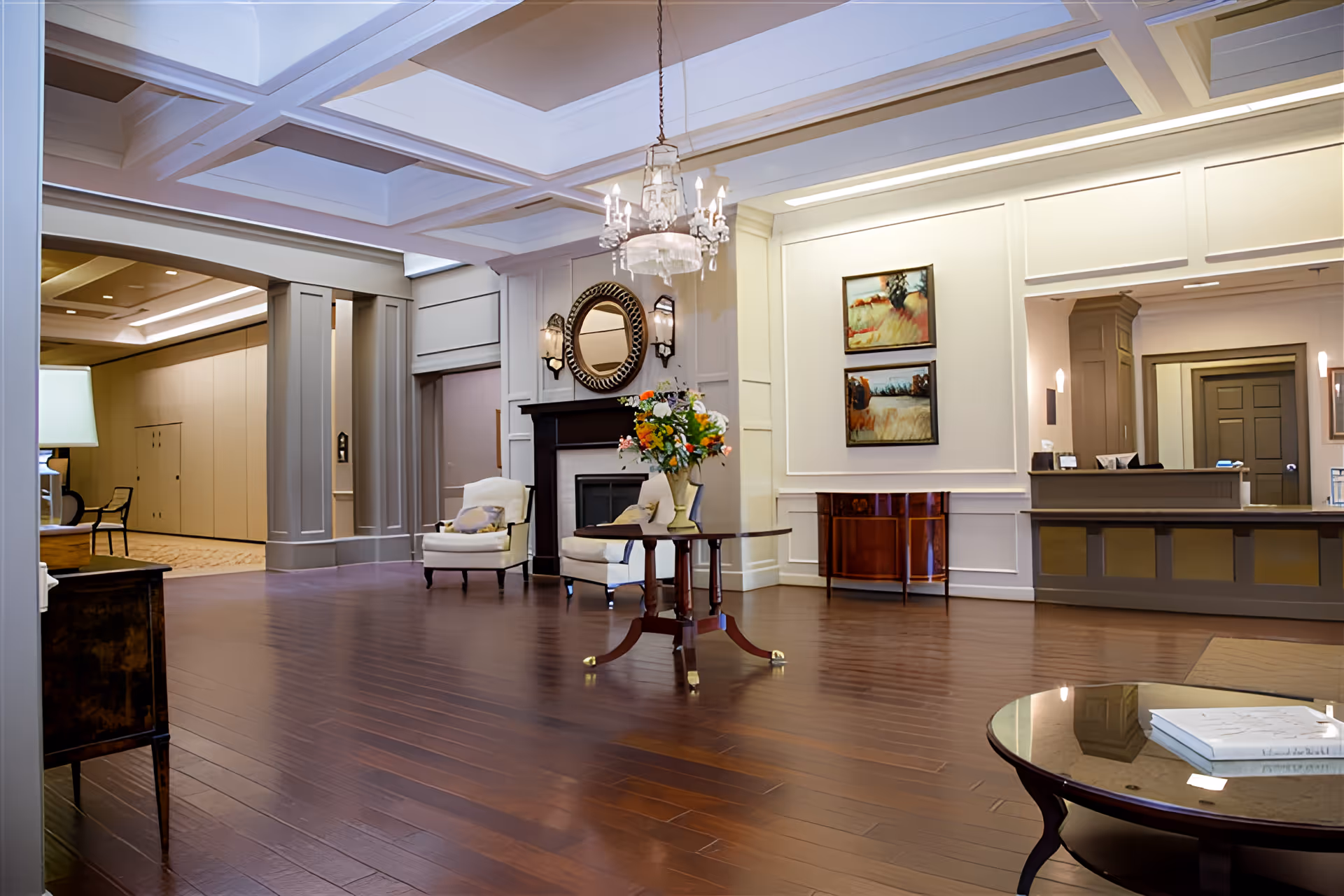 Spacious and elegant senior living facility lobby with polished wooden floors, a central round table with a flower arrangement, two armchairs near a fireplace with a round mirror above it, a chandelier hanging from a coffered ceiling, framed paintings on the wall, and a reception desk in the background.