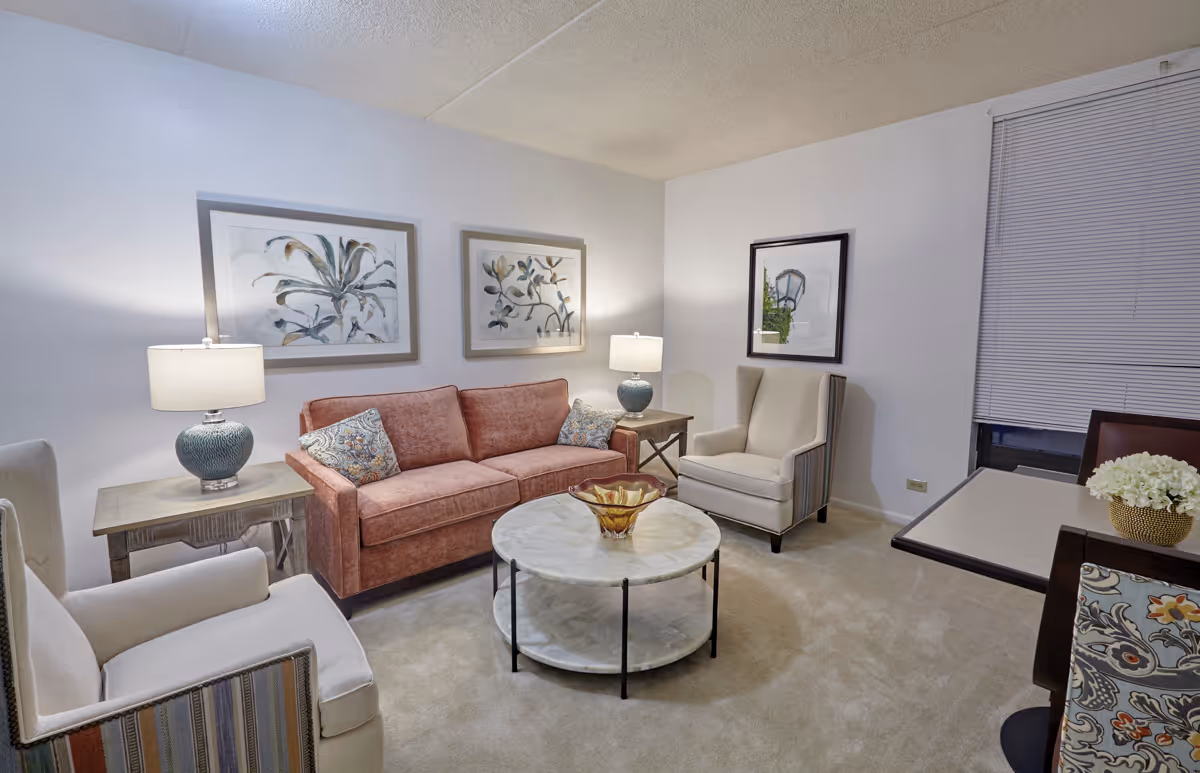 A cozy living room with a pink sofa, two beige armchairs, a round marble coffee table with a decorative bowl, two side tables with lamps, floral artwork on the walls, and a window with closed blinds.