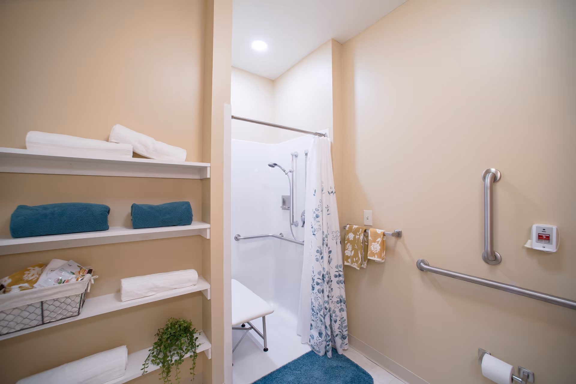 A bathroom with beige walls featuring a walk-in shower with a white shower curtain decorated with blue floral patterns. The shower has a handheld showerhead, grab bars, and a white shower chair. To the left, there are white shelves holding folded white and teal towels, a basket with toiletries, and a small green plant. On the right wall, there are two yellow patterned hand towels hanging on a towel rack, a grab bar, an emergency pull cord, and a toilet paper holder with a roll of toilet paper. A blue bath mat is on the floor in front of the shower.
