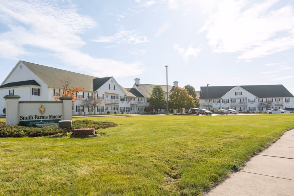 Exterior view of Smith Farms Manor, a large two-story white building with black shutters and a green lawn in front. There is a sign in the foreground that reads 'Smith Farms Manor Five Star Senior Living'. Several cars are parked near the building under a partly cloudy sky.