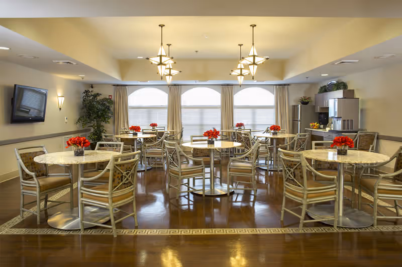 Communal dining room in a senior living facility with round marble-top tables, chairs, pendant lighting, and red floral centerpieces.