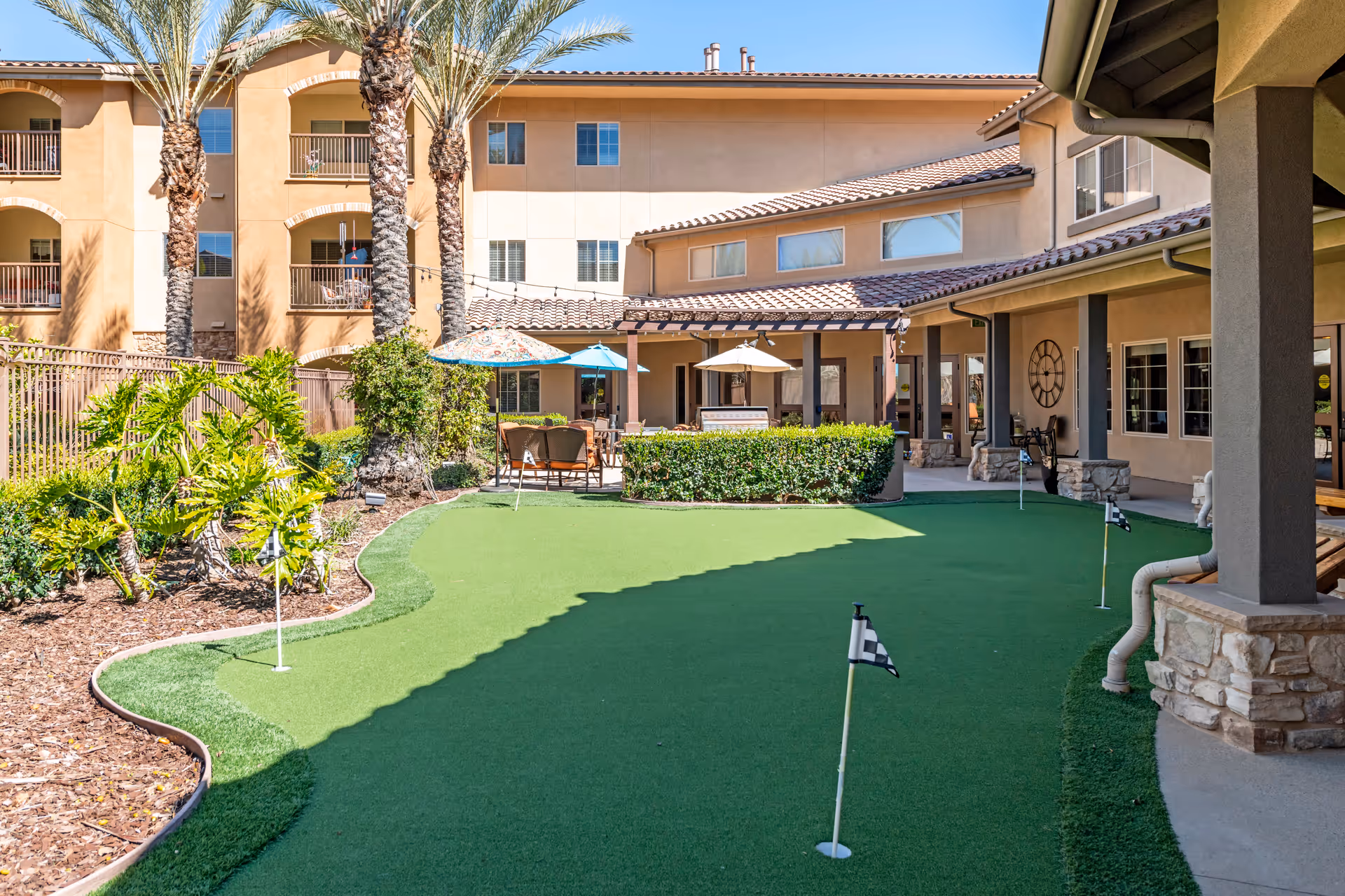 Outdoor putting green area with small golf flags, surrounded by palm trees, plants, and a multi-story building with balconies and shaded seating areas with umbrellas.