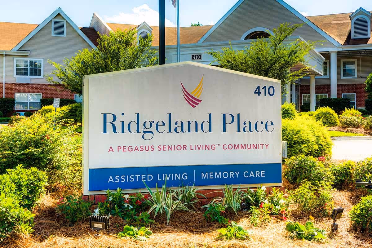 Outdoor view of the Ridgeland Place senior living community sign with the building and landscaping in the background. The sign indicates it is a Pegasus Senior Living community offering assisted living and memory care services.