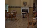 A dining room with several wooden tables and chairs arranged neatly on a tiled floor. There is a fireplace with a framed picture above it on the far wall, and a window with vertical blinds on the left side. The walls are painted beige with a decorative border near the ceiling.
