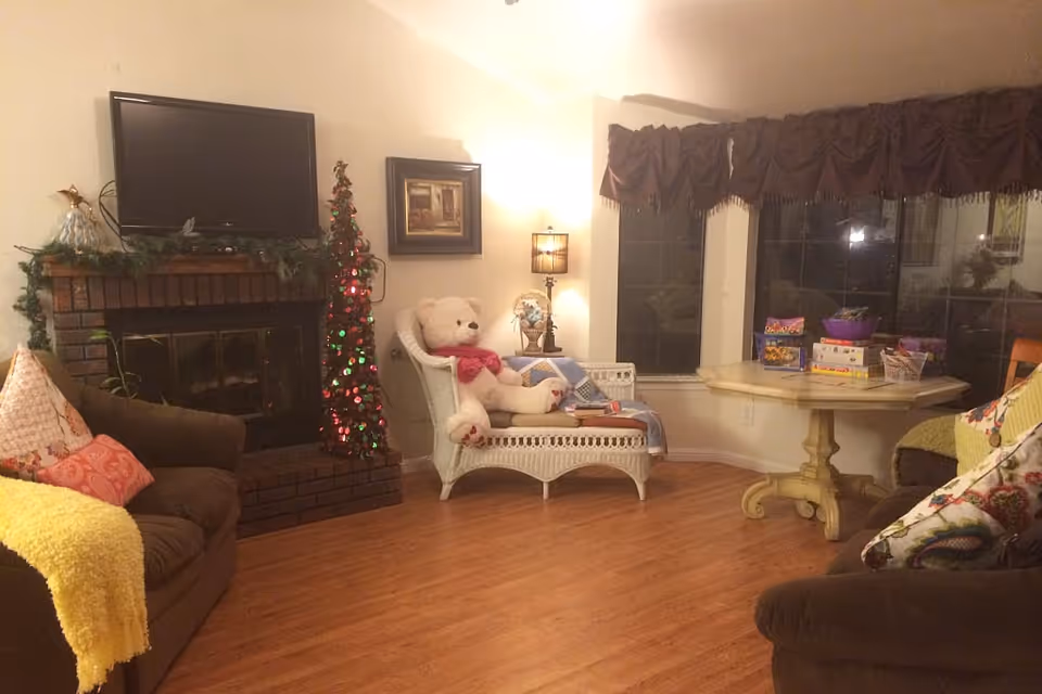 A cozy living room with wooden flooring, a brick fireplace with a TV mounted above it, a small decorated Christmas tree, a white wicker chair with a large teddy bear and a blanket, a round wooden table with board games on it, and two brown sofas with colorful pillows and a yellow throw.