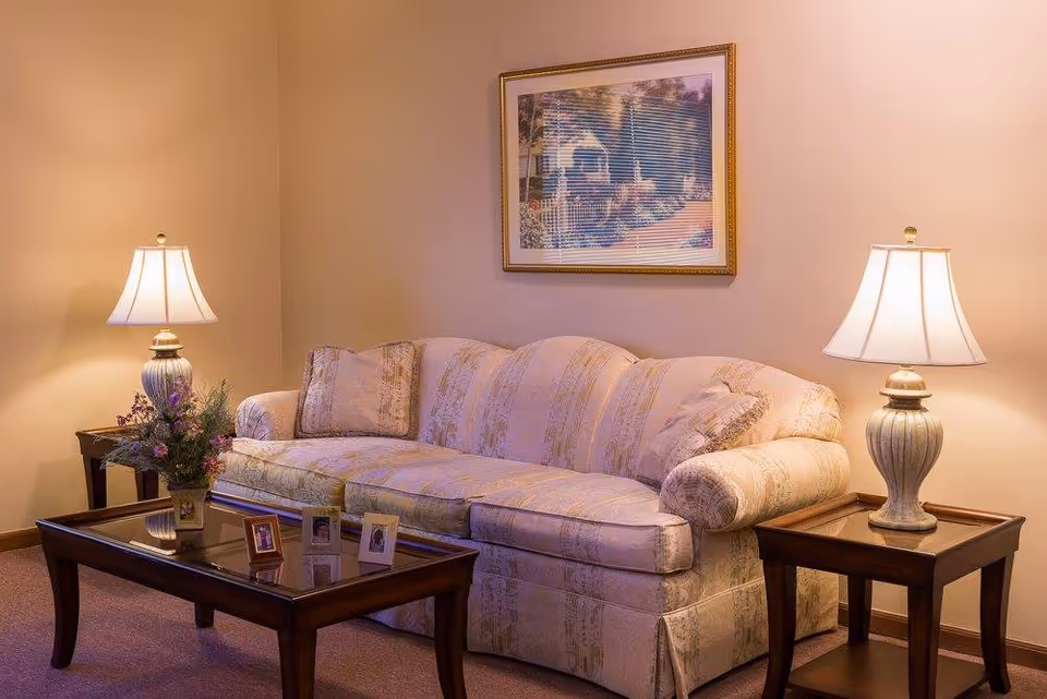 A cozy living room area with a patterned beige sofa, two matching table lamps on wooden side tables, a wooden coffee table with framed photos and a flower arrangement, and a framed painting on the wall.