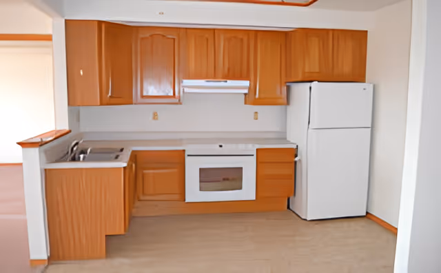 A kitchen area with wooden cabinets, a white refrigerator, a white oven with a range hood, and a sink. The floor is light-colored, and the walls are white. The kitchen is open to an adjacent room with wooden trim.