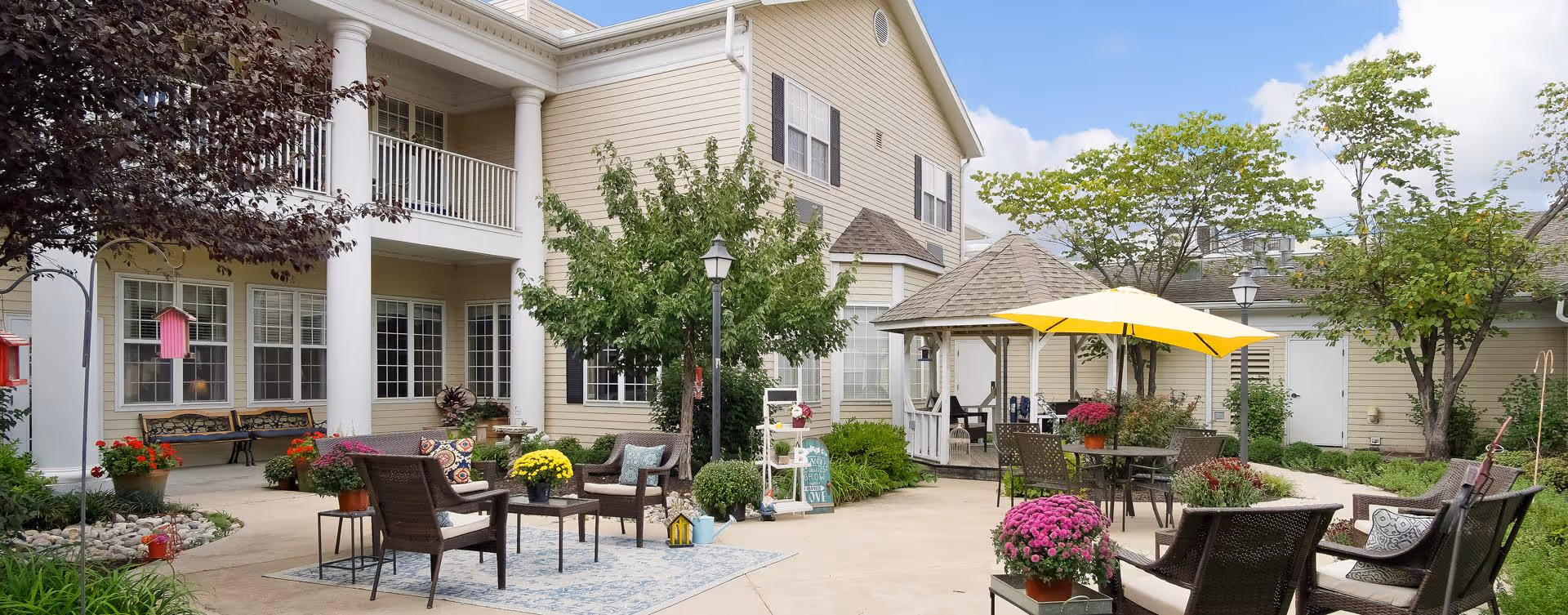 Outdoor courtyard area of a senior living facility with patio furniture, potted flowers, trees, a gazebo with a yellow umbrella, and a beige multi-story building in the background.