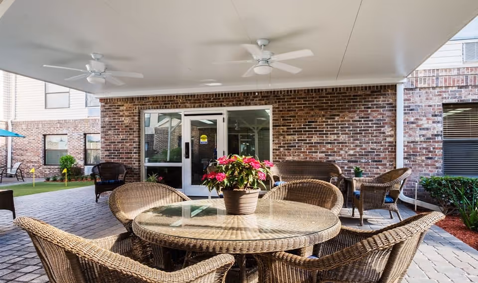 Covered outdoor patio area with a round glass-top wicker table surrounded by wicker chairs. A potted plant with pink flowers is on the table. The patio has ceiling fans and is adjacent to a brick building with glass doors and windows. Additional wicker seating and a small green putting area are visible in the background.