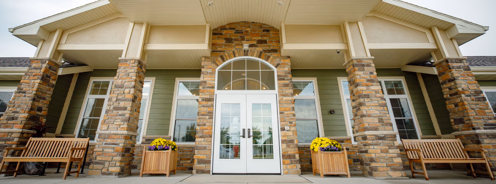 Front entrance of a senior living building with stone columns, a double glass door with an arched transom, benches, and flower planters.
