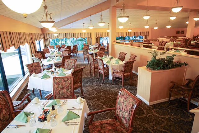 A spacious dining room with multiple tables covered in white tablecloths and set with green folded napkins, glasses, and silverware. The room features patterned carpet, floral upholstered chairs, large windows with striped valances, and hanging ceiling lights.
