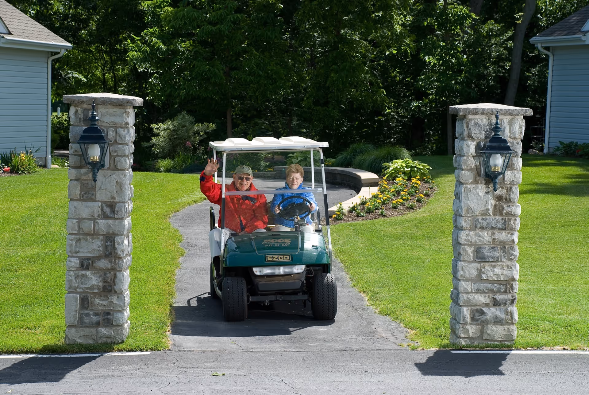 Two elderly people riding a green golf cart on a paved path between two stone pillars with lanterns, surrounded by green grass and trees in a sunny outdoor setting.