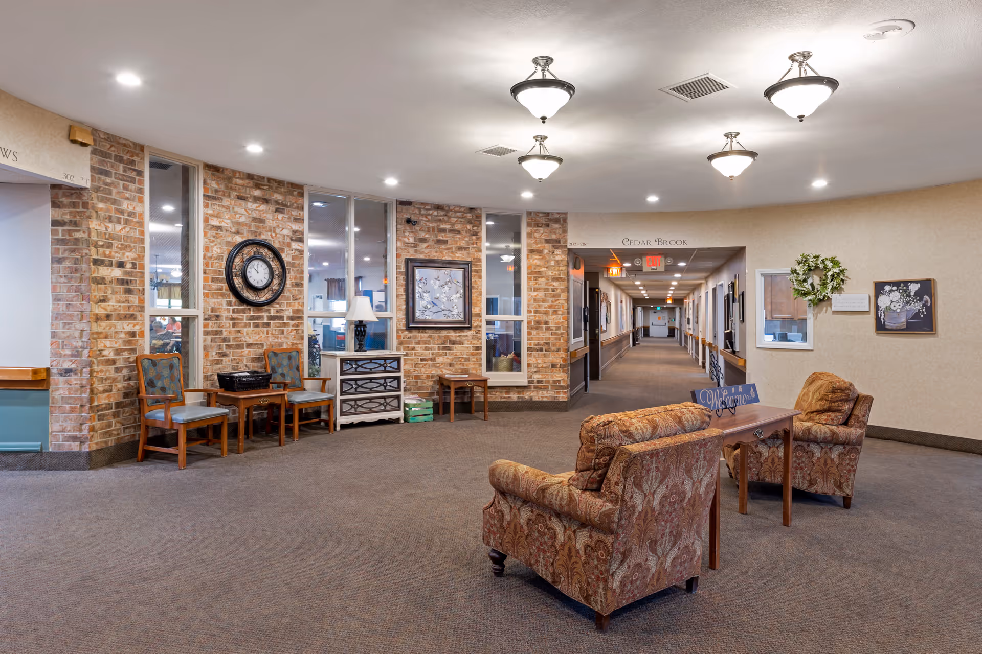 Spacious rehabilitation center lobby with patterned armchairs and small tables in the foreground, a brick accent wall with chairs and a clock to the left, and a long corridor leading to rooms in the background.