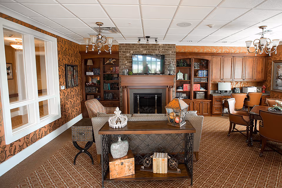 A cozy living room area with patterned carpet and wallpaper, featuring a fireplace with a TV mounted above it, built-in wooden bookshelves filled with books and decorative items, a sofa facing the fireplace, a wooden table behind the sofa with decorative objects, and a dining table with chairs to the right. There is also a desk area with a computer and printer in the background.