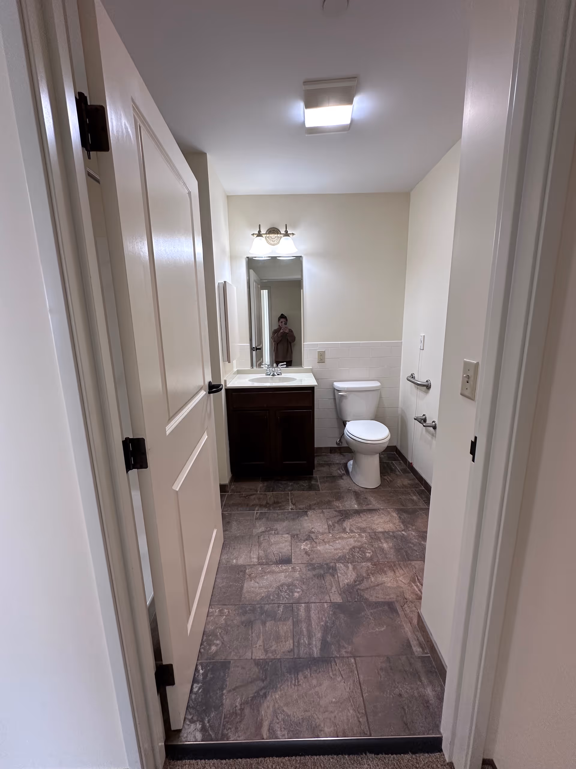 View into a bathroom with a white door partially open, showing a toilet, a sink with a dark wood cabinet, a mirror above the sink, and a light fixture above the mirror. The floor has brown and gray tiles, and the walls are light-colored.