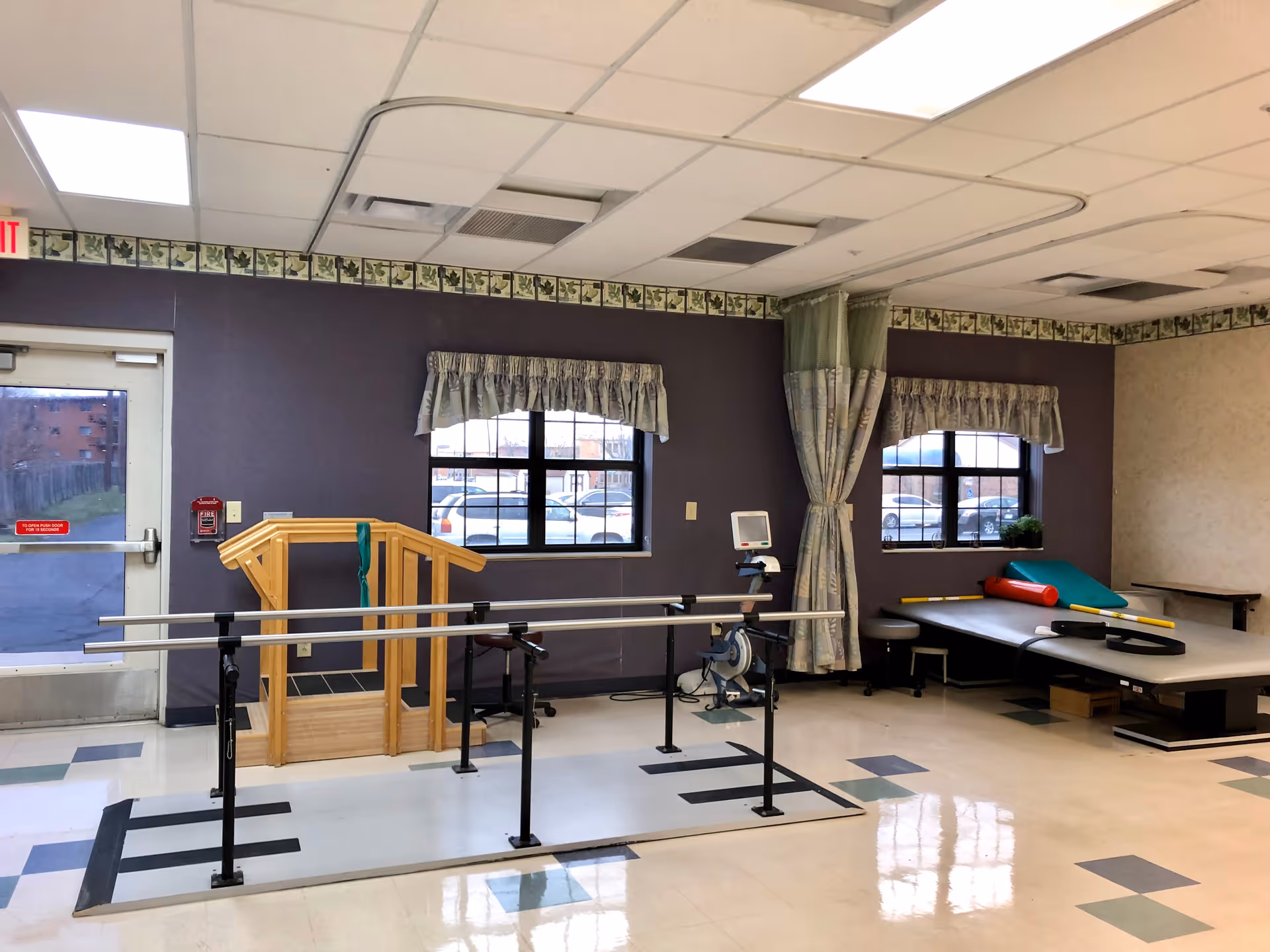 A rehabilitation room with parallel bars for walking exercises, a wooden ramp with handrails, a stationary exercise bike, and a padded therapy table with various cushions and supports. The room has two windows with curtains, a door leading outside, and a tiled floor with a patterned border near the ceiling.