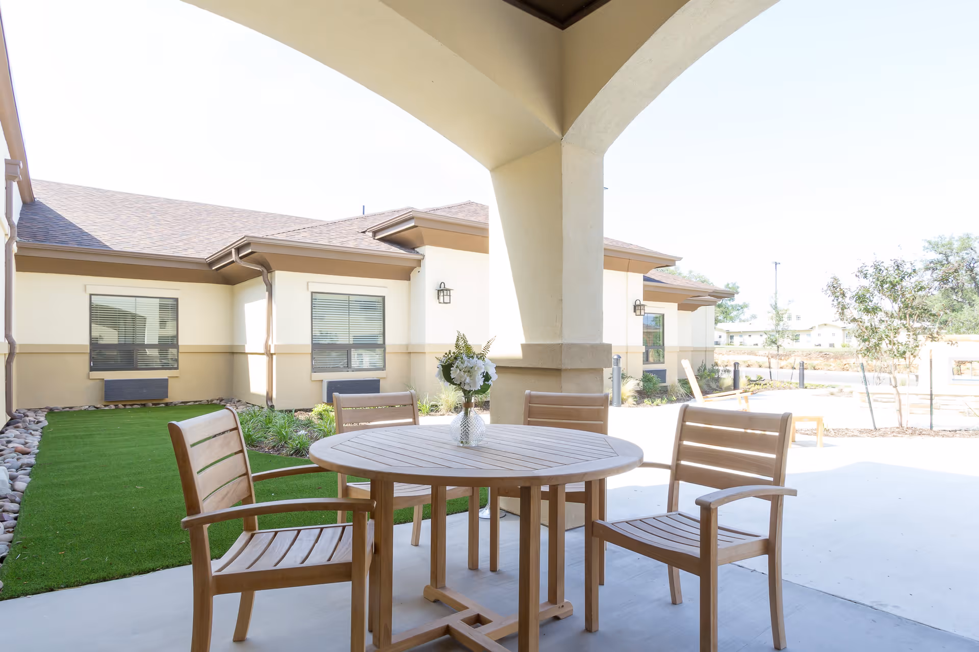 Outdoor covered patio area with a round wooden table and four wooden chairs. A vase with white flowers is placed on the table. The patio overlooks a well-maintained lawn and the exterior of a beige building with windows and brown trim.