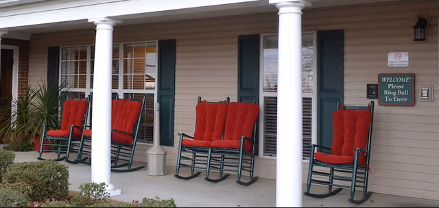 Covered front porch with white columns and several green rocking chairs with red cushions outside the entrance.