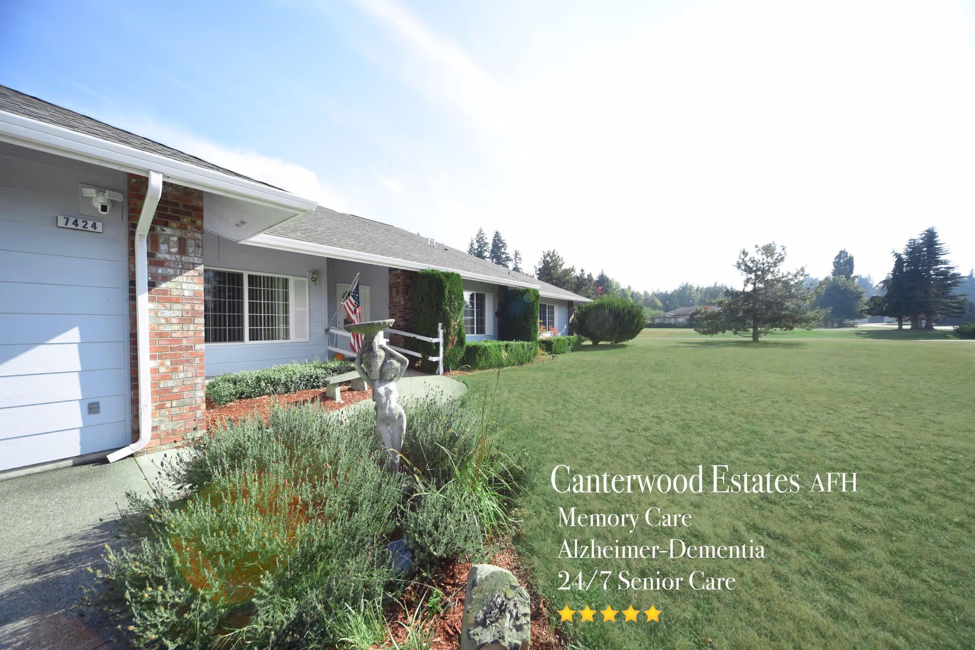 Exterior view of Canterwood Estates Adult Family Home showing a single-story building with brick and light blue siding, a well-maintained lawn, shrubs, a small statue, and an American flag near the entrance under a clear blue sky.