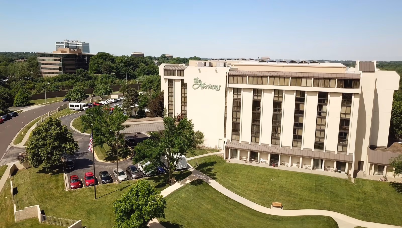 Aerial view of The Atriums Senior Living Community building surrounded by green lawns, trees, and a parking lot with several cars. The building is multi-story with large windows and a sign displaying 'The Atriums' on the side. A curved walkway and an American flag are visible in the foreground.