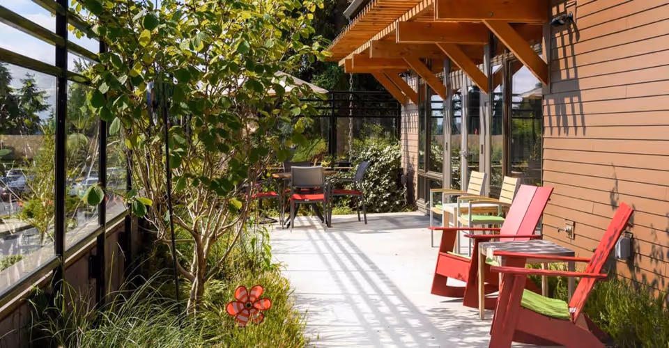 Outdoor patio area with red and beige chairs, a small table, and a tree surrounded by plants. The patio is adjacent to a building with large windows and wooden siding, and is enclosed by glass panels.