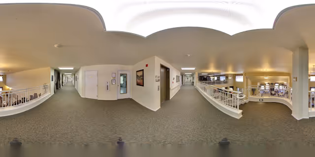 Carpeted interior corridor and landing in a senior living facility overlooking an open atrium with white railings and seating below.