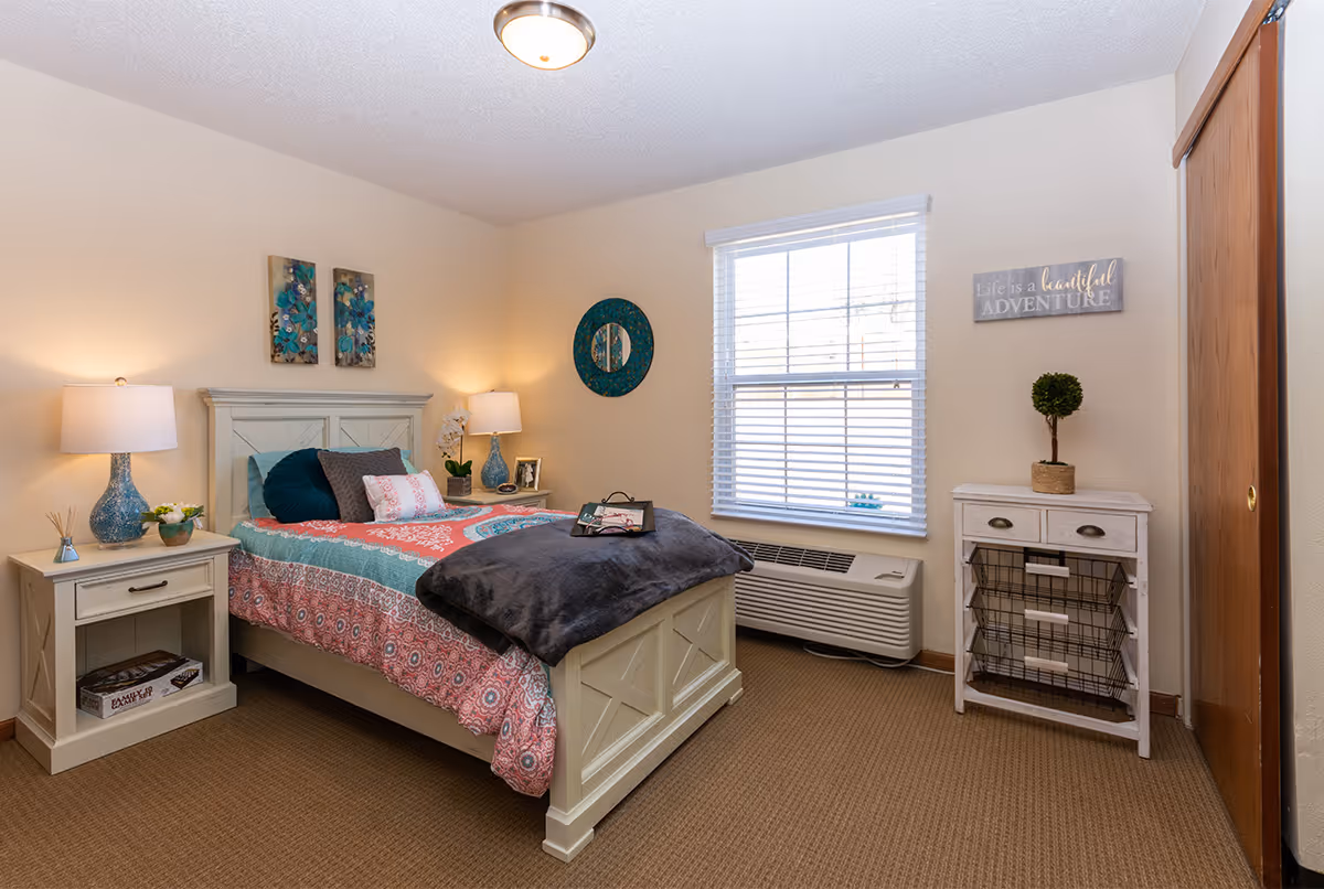 A cozy bedroom with a single bed featuring a colorful quilt and multiple pillows. There are two bedside tables with lamps and decorative items, a window with blinds, a small white cabinet with baskets, and wall decorations including a round mirror and a sign that reads 'Life is a beautiful ADVENTURE'.