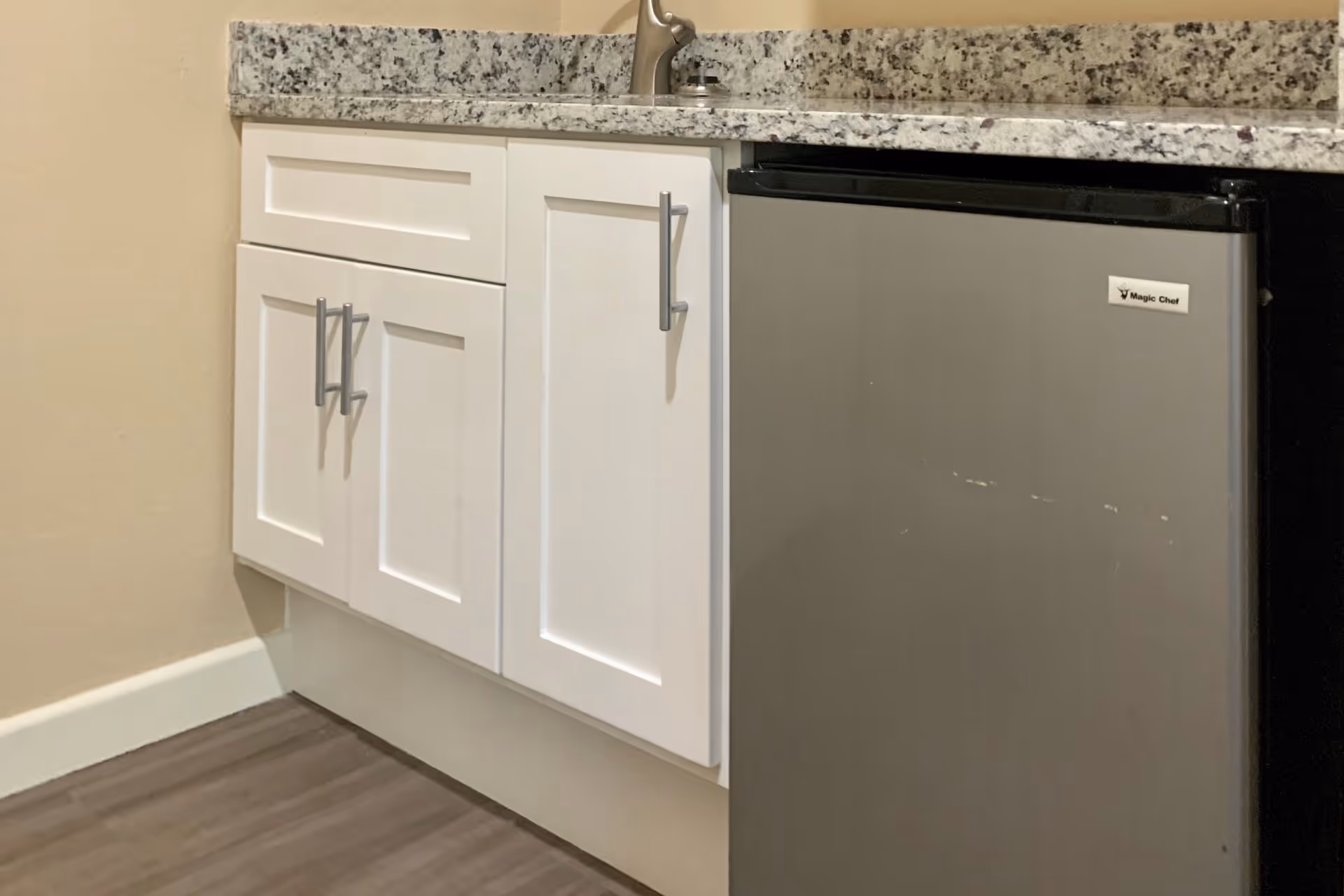 A close-up view of a kitchen area featuring white cabinets with silver handles, a granite countertop with a faucet, and a small stainless steel Magic Chef refrigerator. The floor is wood-style laminate and the walls are painted beige.