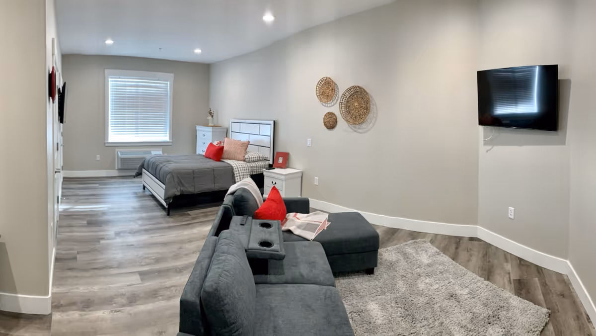 A modern, well-lit room with a bed featuring gray bedding and red accent pillows, a white nightstand and dresser, a gray sectional sofa with red pillows, a wall-mounted flat-screen TV, and decorative woven wall hangings. The floor is wood with a light gray area rug.