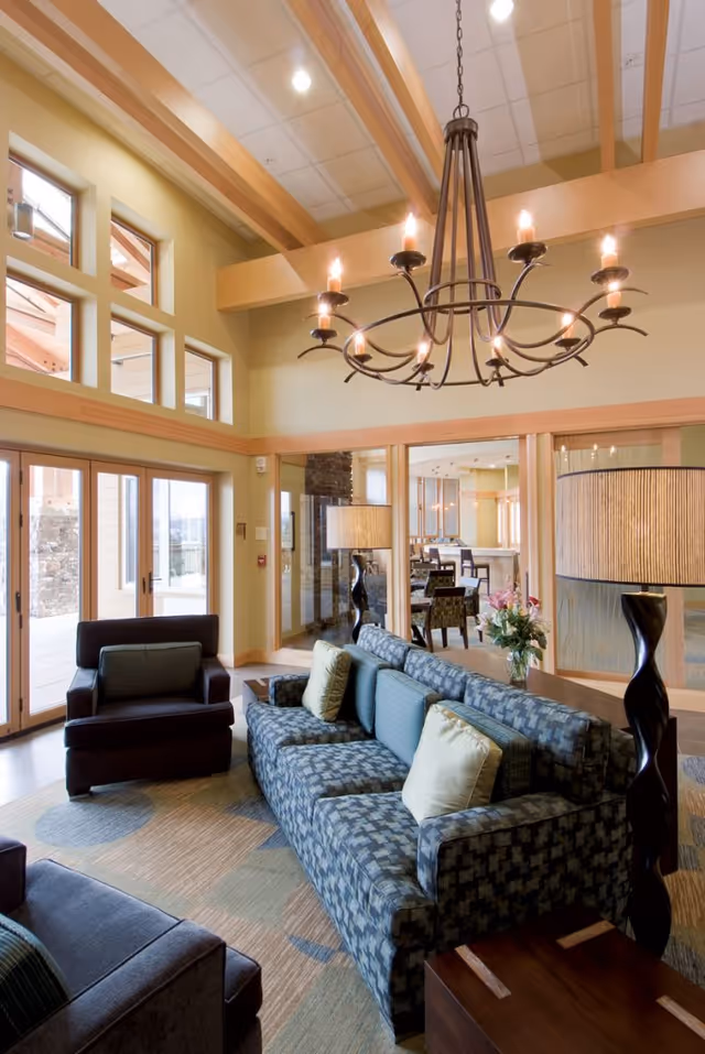 Sunlit common lounge with a patterned sofa and armchairs, a chandelier, large windows, and a view into a dining area.