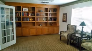 A cozy interior room with a large wooden built-in bookshelf filled with books and decorative items. Two upholstered armchairs are positioned near a window with blinds, accompanied by a small side table with a lamp. The room has carpeted flooring and a glass-paneled door on the left side.