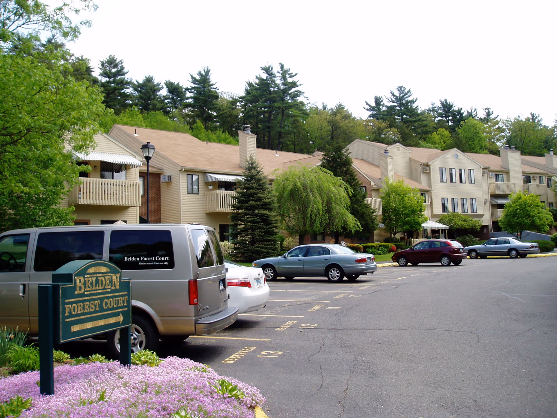 Exterior view of Belden Forest Court Inc, showing a parking lot with several parked cars and a van with the facility's name on the side. The building is a two-story residential structure with balconies, surrounded by trees and greenery.