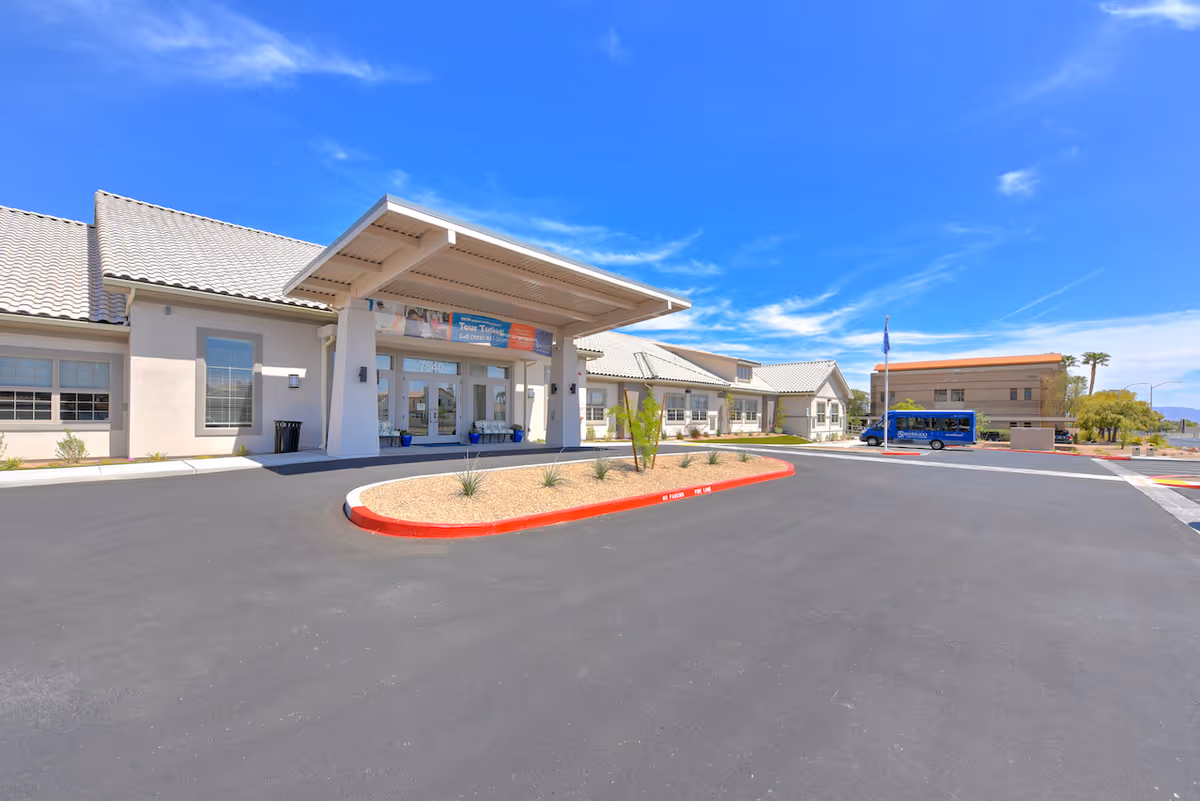 Front entrance of a single-story memory care facility with a covered drop-off, circular driveway and a blue shuttle bus under a bright blue sky.