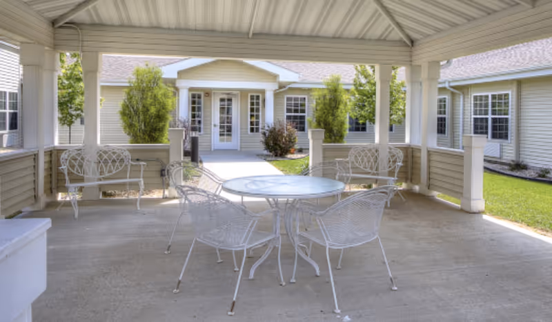 Covered courtyard patio with a round white metal table and matching chairs facing the entrance of the senior living building.