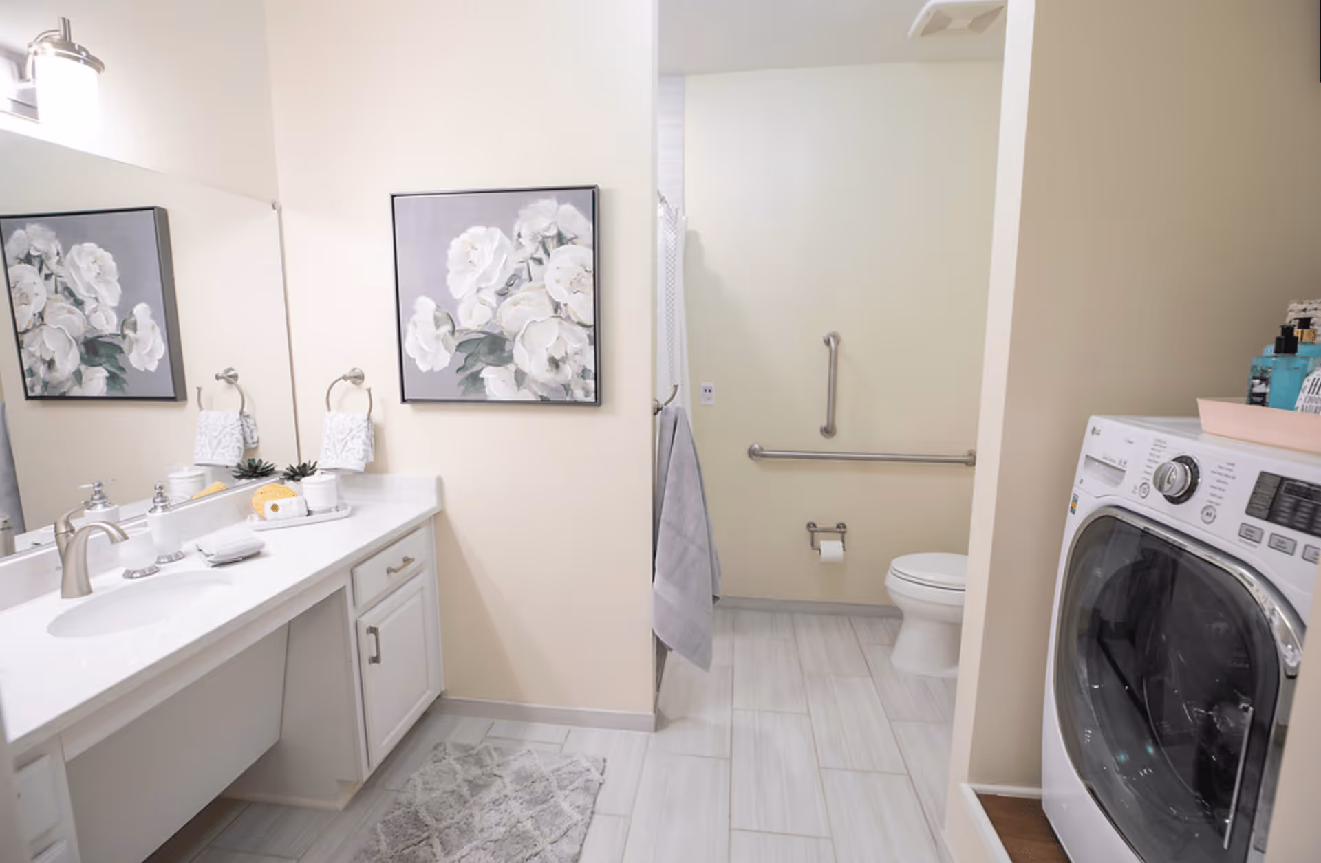 A clean and modern bathroom with a white vanity and sink on the left, a large mirror above it, and a floral painting on the wall. The bathroom includes a toilet with grab bars for accessibility and a shower area partially visible. On the right side, there is a front-loading washing machine with various toiletries on top. The floor is tiled in light gray, and a gray bath mat is placed in front of the vanity.