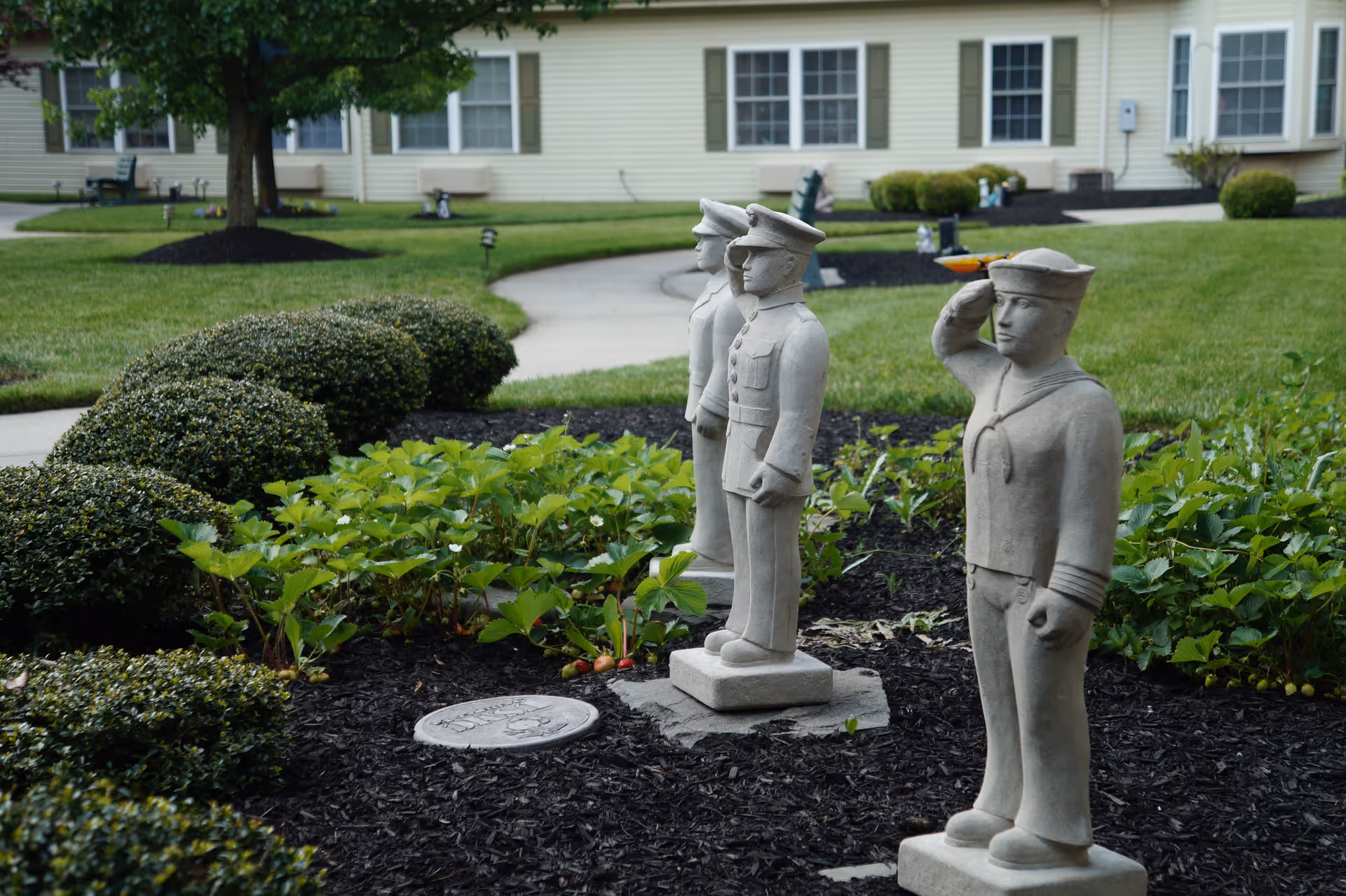 Three stone statues of military personnel, including a sailor saluting, standing in a landscaped garden area with bushes and plants. A curved concrete pathway and a beige building with windows are visible in the background.