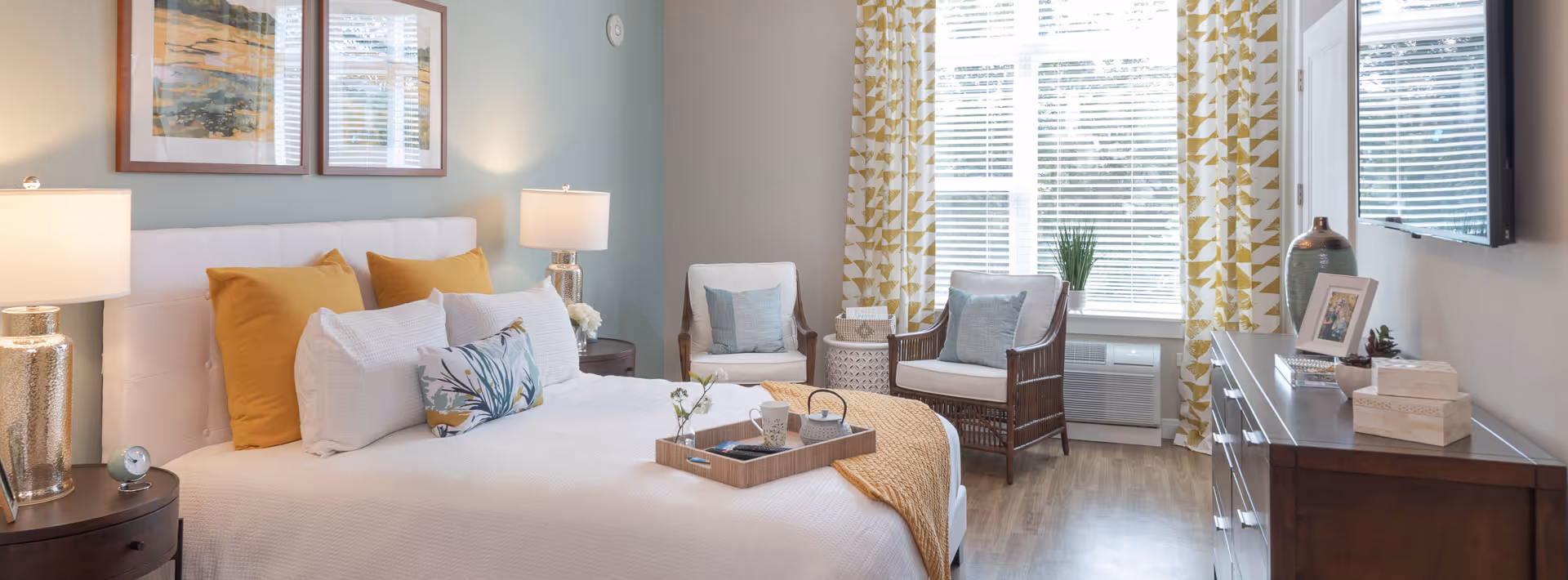 A bright and cozy bedroom with a large bed featuring white and mustard yellow pillows, a white headboard, two bedside tables with lamps, two wicker chairs with cushions near a window with yellow patterned curtains, a wooden dresser with decorative items, and a wall-mounted TV.