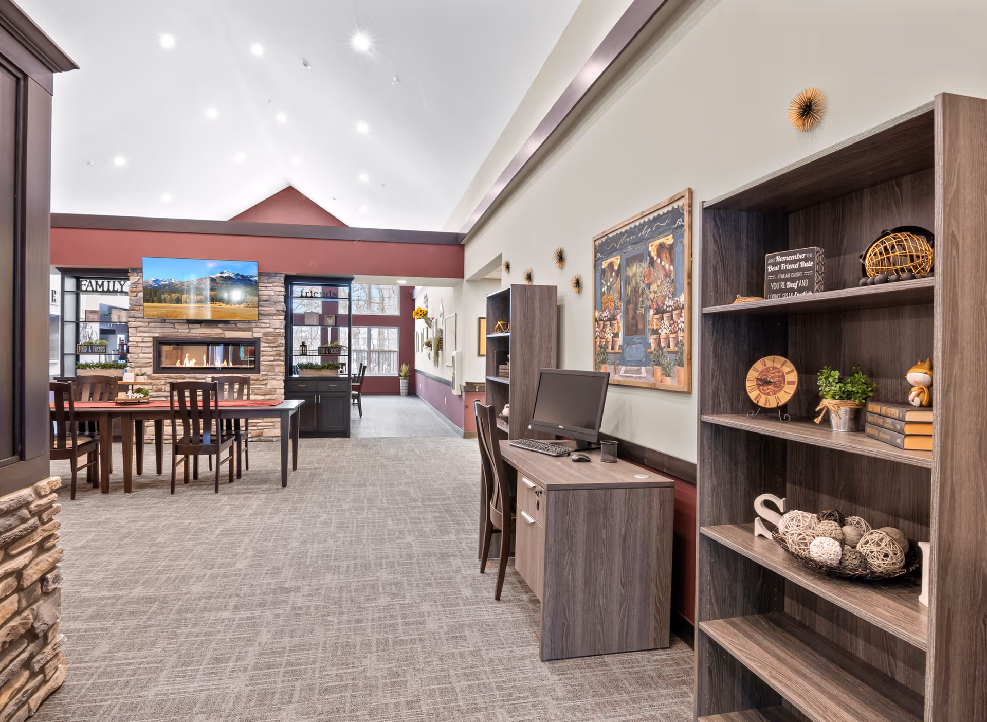 A bright and spacious common area in a senior living facility featuring a stone fireplace with a mounted flat-screen TV above it. There are wooden tables and chairs arranged for dining or socializing. On the right side, there is a wooden desk with a computer and a matching bookshelf decorated with various ornaments and books. The walls are adorned with artwork and decorative wall hangings, and large windows let in natural light.