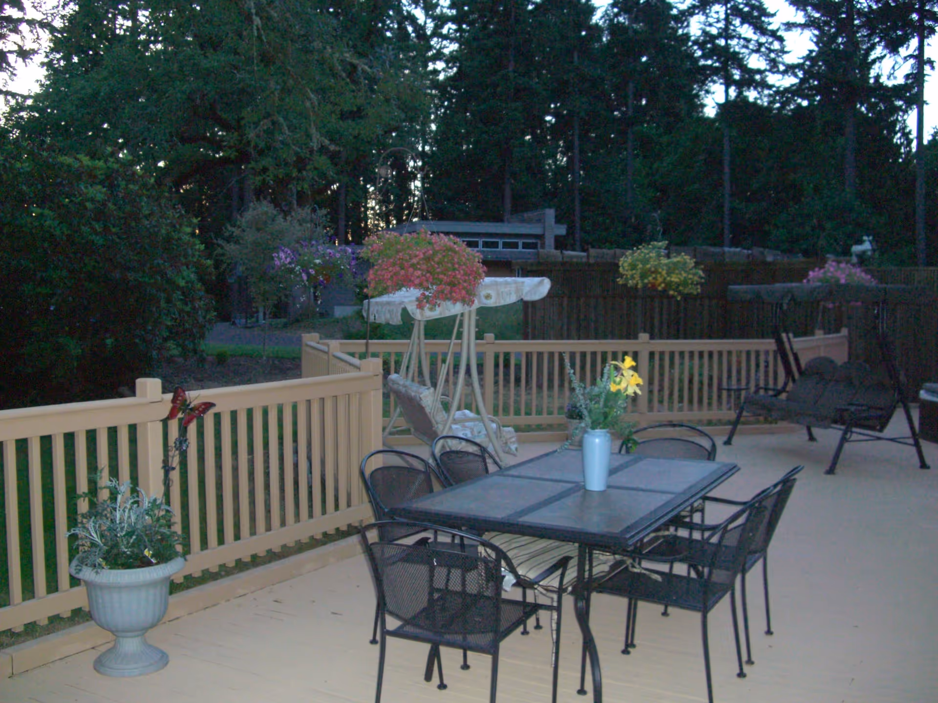 Outdoor patio area with a metal table and six chairs, a vase with yellow flowers on the table, a cushioned swing with a canopy, hanging flower baskets, and surrounding trees and greenery.