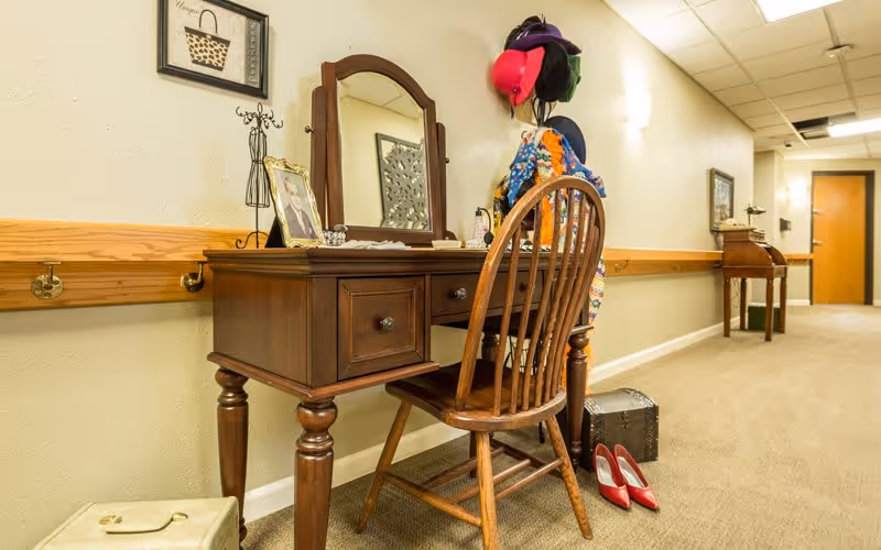 A hallway in a senior living facility with a wooden vanity table and chair placed against the wall. The vanity has a mirror, framed photo, and decorative items on top. A coat rack next to the vanity holds colorful hats and scarves. On the floor near the vanity are a small suitcase and a pair of red high-heeled shoes. The hallway is carpeted and has handrails along the walls, with doors visible in the background.